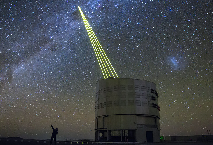 A watchtower in the desert that connects to a satellite at night. Next to it is a man with a camera