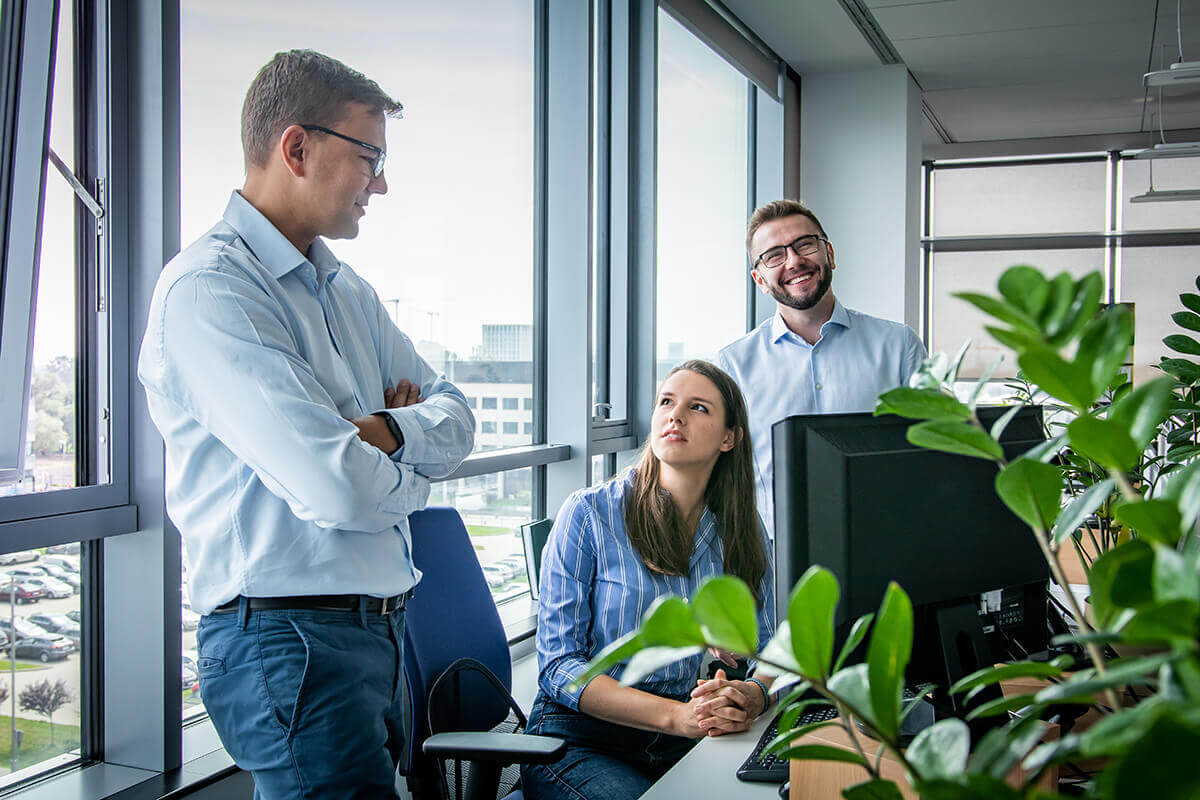 Three Comarch employees talk to each other about the project. Two men stand and a woman sits