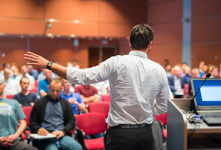 The man at the ScalaSphere DevTools Summit. In the background, there are men sitting down