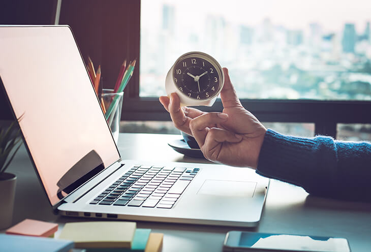 The man is holding a clock while working on a laptop. He wonders how to manage working time