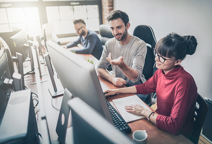 A young man explains something to his work colleague while working on a computer