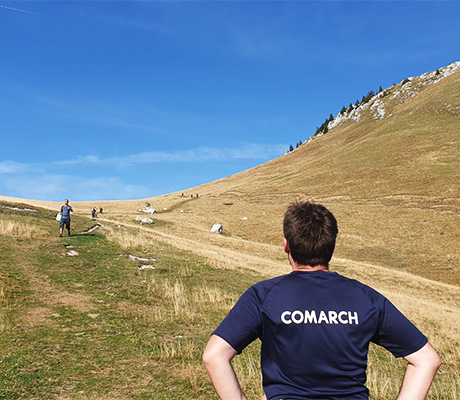 The man standing on the beach at the seaside, there are mountains all around, and he's showing on a T-shirt with a Comrach logo