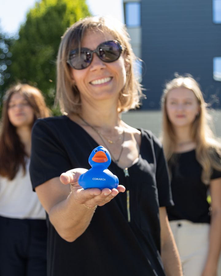 smiling woman holding a blue rubber duck branded with the Comarch logo, with colleagues and an office building in the blurred background.