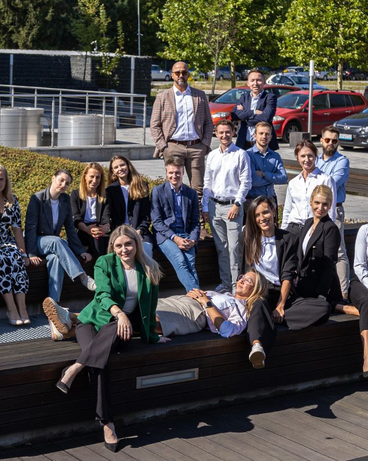A diverse group of Comarch employees in business casual attire posing together outdoors in a relaxed, friendly atmosphere.